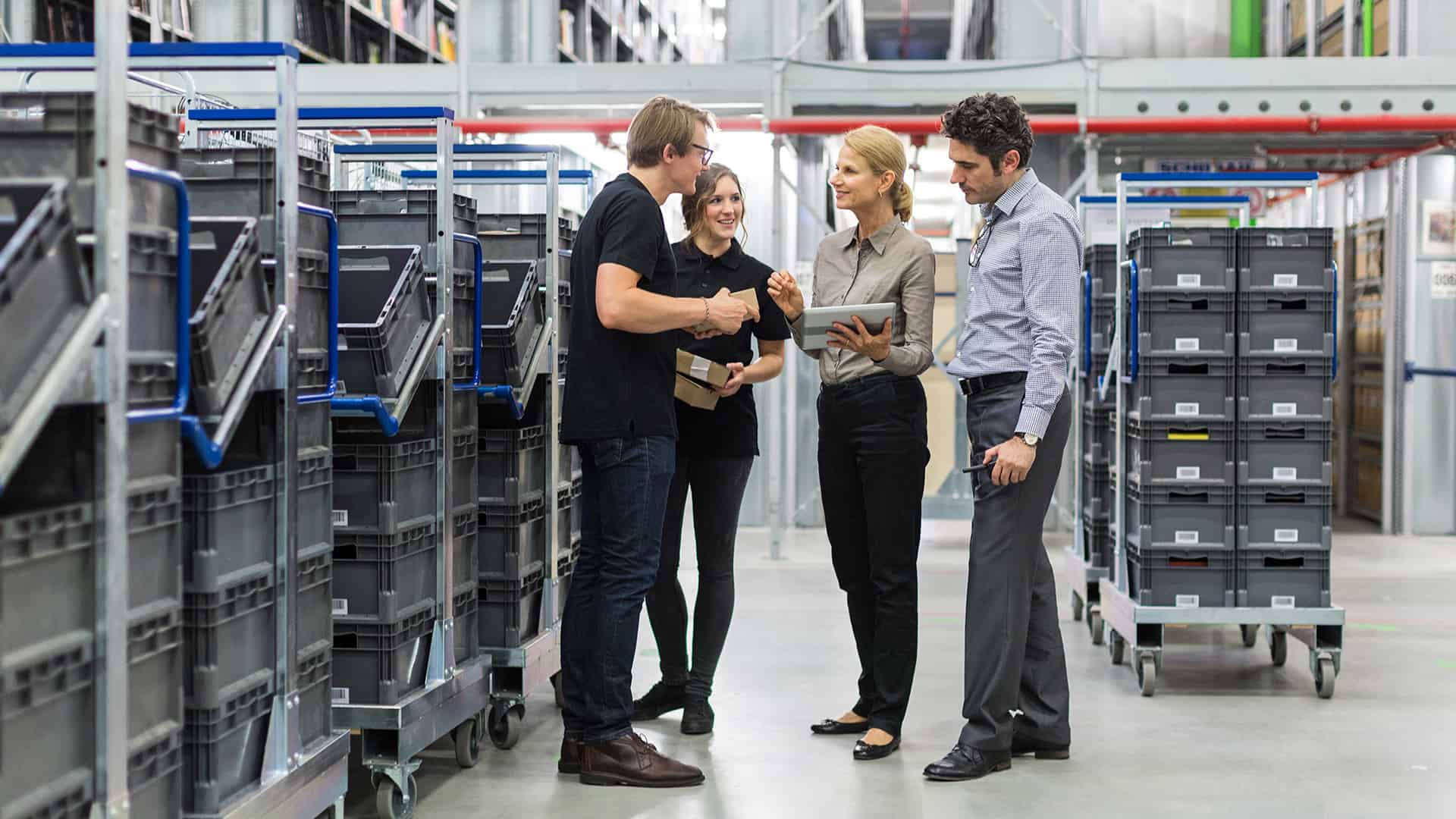 Warehouse team discussing logistics with boxes and trolleys in the background.
