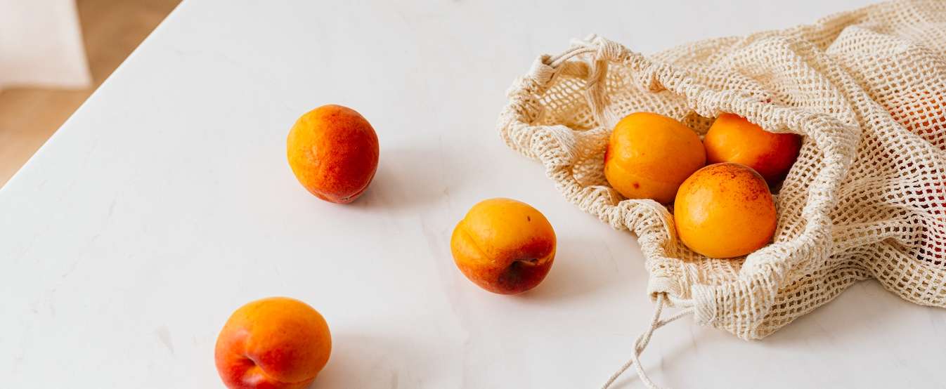 Fresh apricots spilling from a reusable mesh bag on a white surface, showcasing eco-friendly shopping habits.