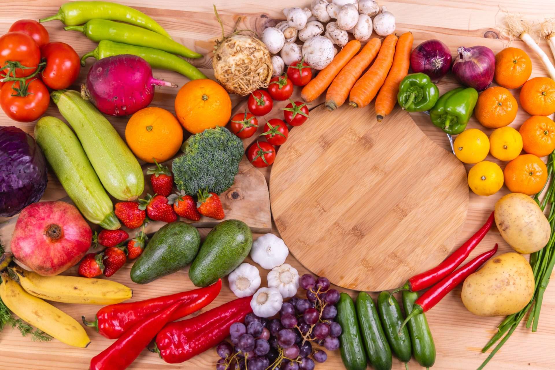 Assorted fresh fruits and vegetables arranged around an empty wooden cutting board on a kitchen table.